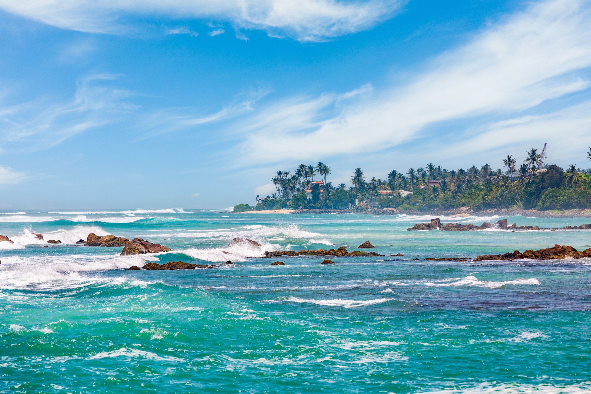 Turquoise Ocean Waves Crashing on Rocky Shoreline, Lush Green Coastal Landscape Under Blue Sky with Wispy Clouds.